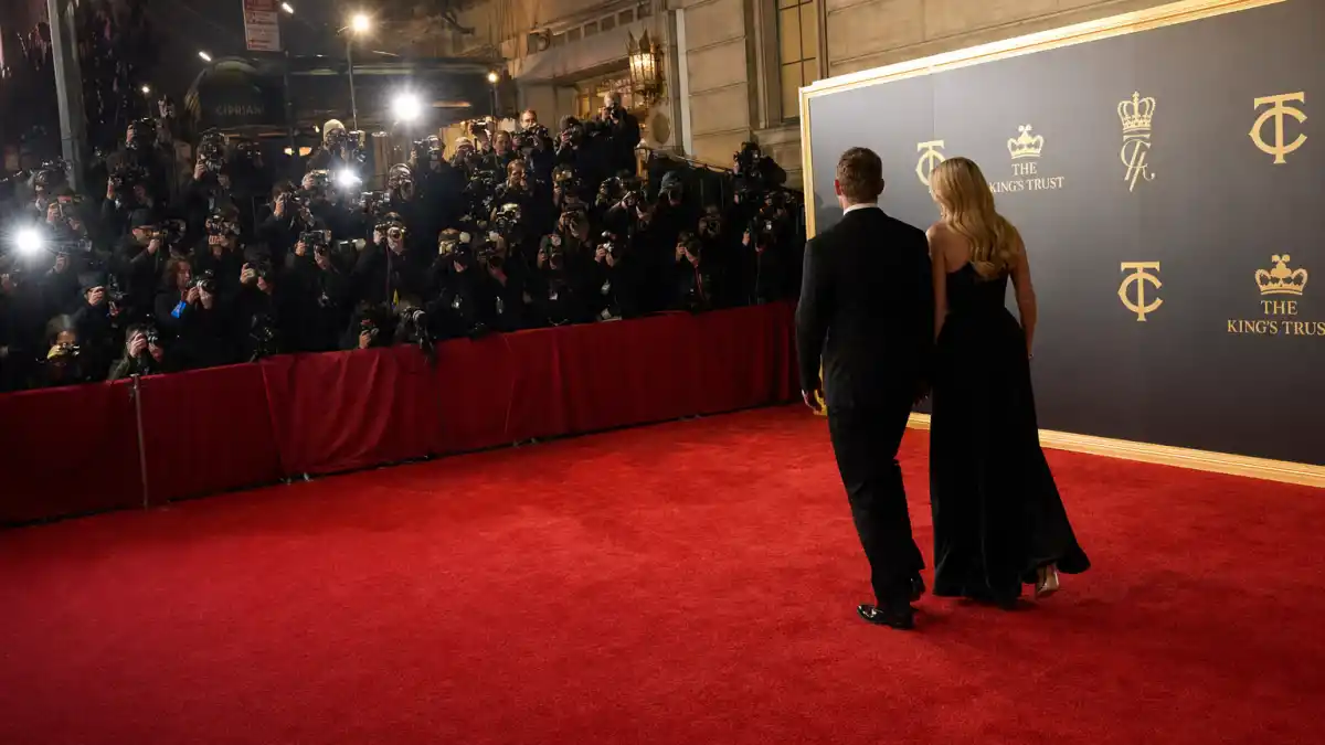 A formally-dressed couple seen from behind walking down the red carpet at the King's Trust Gala 2026 in New York, with paparazzi flashes lighting the step-and-repeat backdrop
