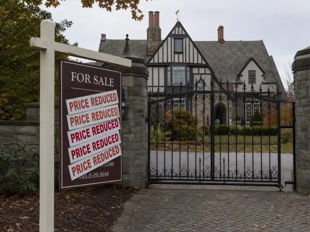 Layered price reduction stickers on a luxury real estate listing sign for celebrities who can't sell their mansion