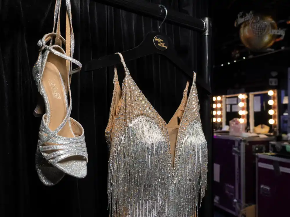 Glittery silver Latin ballroom dance shoe and sequined fringe gown backstage at a BBC Strictly Come Dancing-style set where Tess Daly hosts