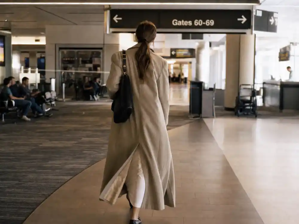 Anonymous figure from behind in airport lounge wearing cream slip dress and longline trench coat illustrating 90s supermodel off duty style trend airport glamour