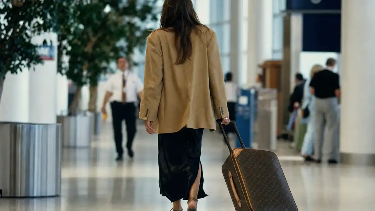 90s supermodel off duty style trend — anonymous woman from behind in black silk slip dress and oversized camel blazer walking through airport terminal with rolling suitcase
