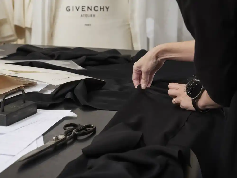 Black silk crepe draped on couture cutting table with seamstress hands adjusting hem behind Audrey Hepburn little black dress Givenchy construction process
