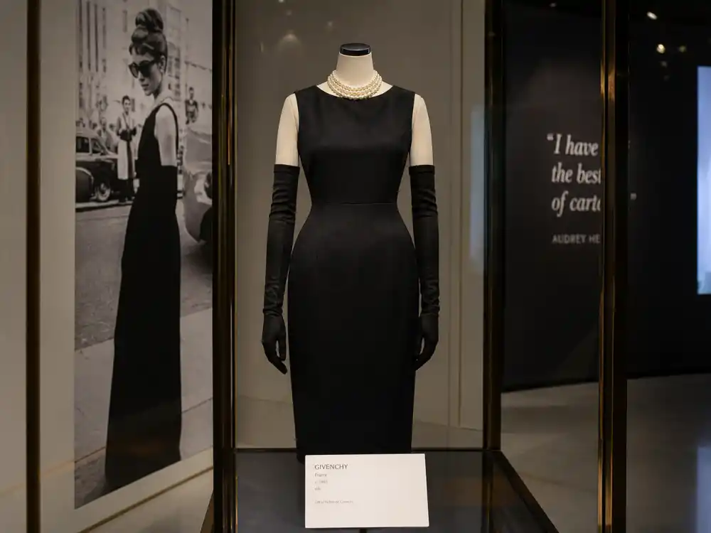 Fashion museum glass case preserving Audrey Hepburn little black dress Givenchy legacy — black sheath gown with pearls on gallery mannequin under spotlights
