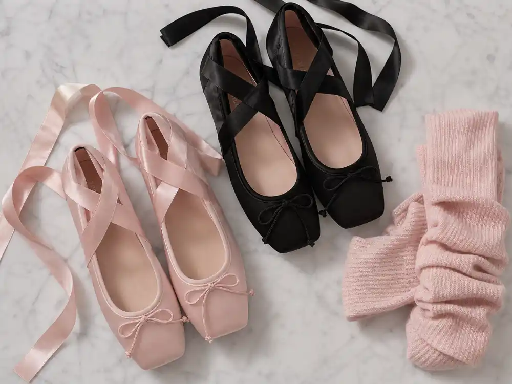 Blush pink and black satin ballet flats with ribbon ties beside soft leg warmers flat lay for balletcore celebrity fashion trend footwear foundation