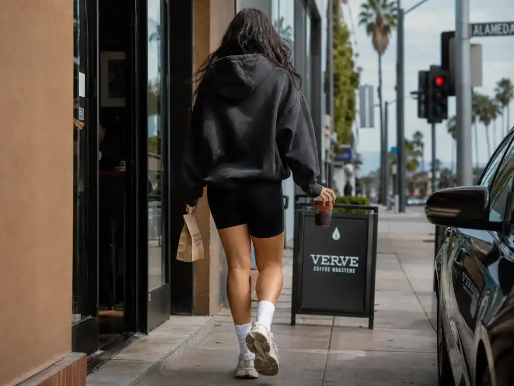 Coffee-run exit with anonymous woman from behind in oversized hoodie, bike shorts, and chunky sneakers holding iced coffee for best celebrity street style of all time Los Angeles errands