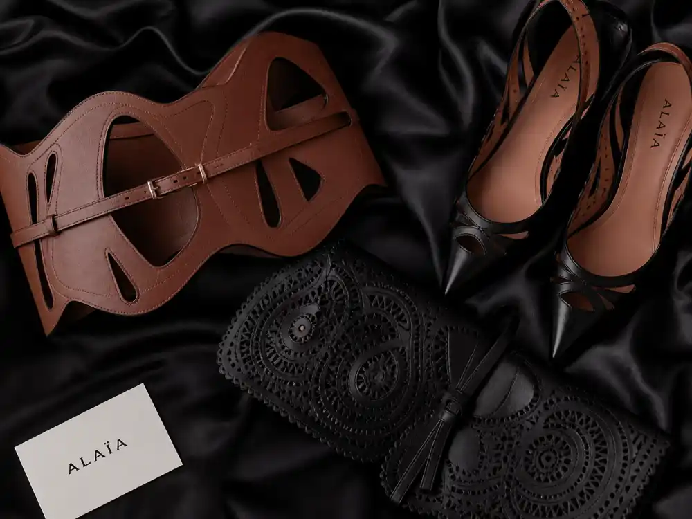 Alaïa sculptural leather belt laser-cut clutch and pointed heels flat lay on black silk capturing accessory finishing rules celebrities who wear Alaia layer with bodycon knit columns