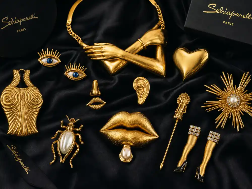 Schiaparelli gold surreal body jewelry pieces and sculptural brooches flat lay on black silk capturing accessory stacking rules celebrities who wear Schiaparelli use when couture alone cannot carry camera