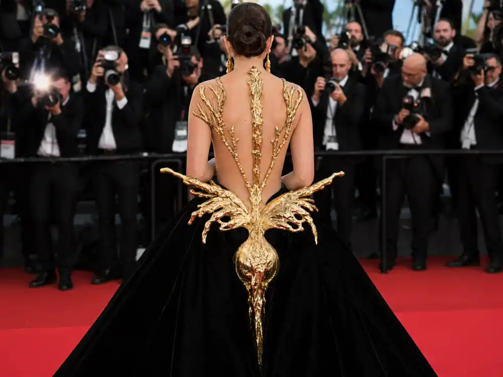 Anonymous woman photographed from behind in dramatic Schiaparelli avant-garde evening gown with gold sculptural details at step-and-repeat showing how celebrities who wear Schiaparelli command red carpet visibility
