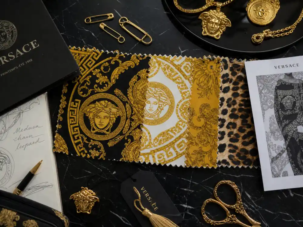 Baroque print fabric swatches gold safety pins and Medusa jewelry flat lay capturing the print language celebrities who wear Versace layer across gowns and tailoring