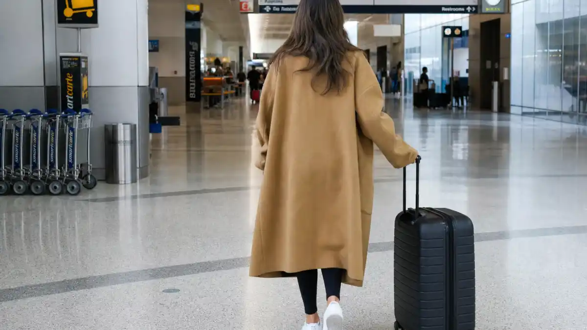 Celebrity airport fashion street style — camel wool coat black leggings white sneakers and rolling luggage at modern airport terminal seen from behind