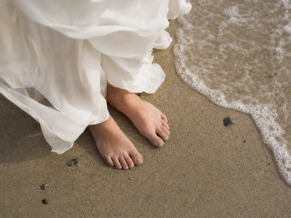 Barefoot bride hem swirling in sand and wave foam completing the accessory formula behind iconic celebrity beach wedding dress looks