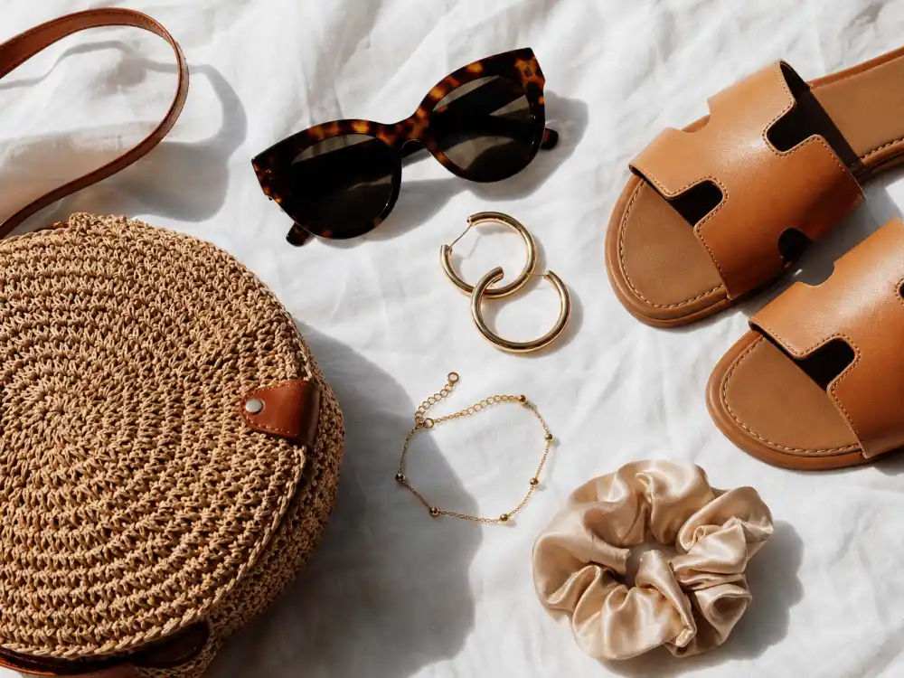 Cat-eye sunglasses gold hoop earrings woven straw crossbody bag tan leather slide sandals and silk scrunchie flat lay completing accessory stacking rules for celebrity brunch outfit ideas finishing touches