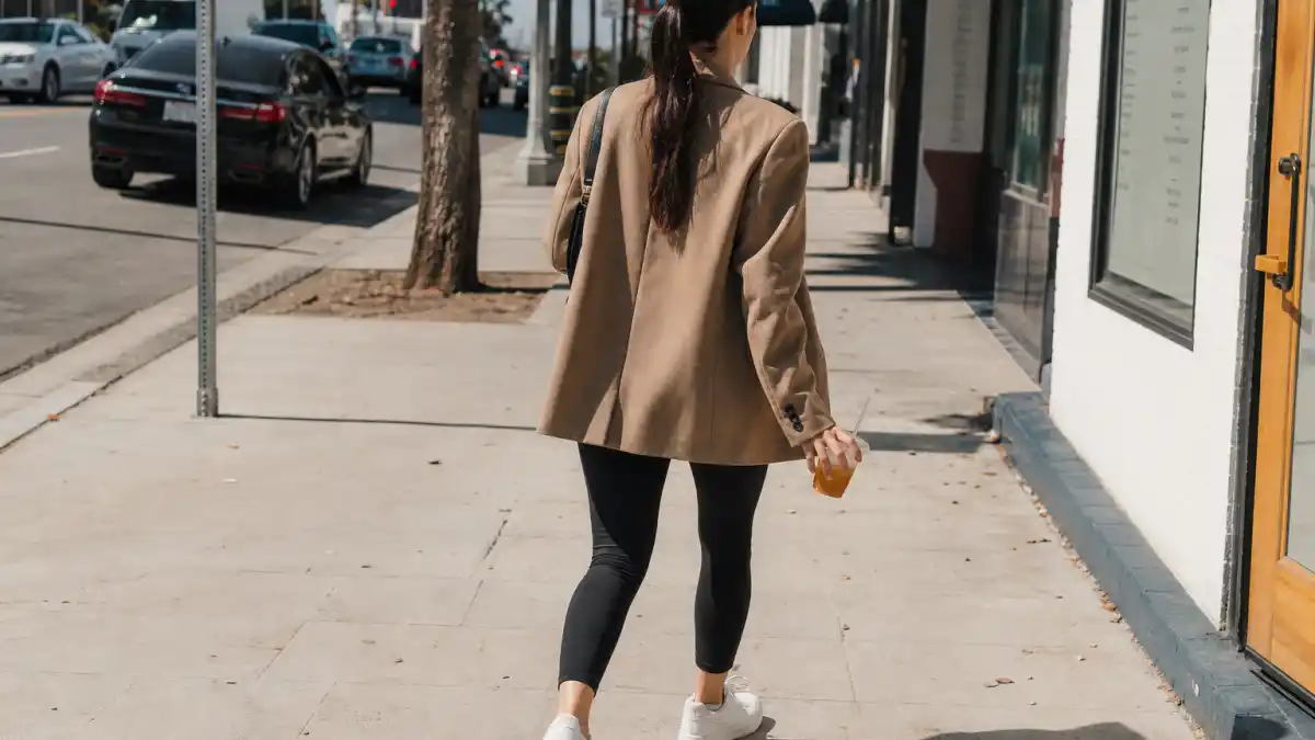 Celebrity casual errands style 2026 — camel oversized blazer black leggings white sneakers and iced coffee on sunny city sidewalk seen from behind