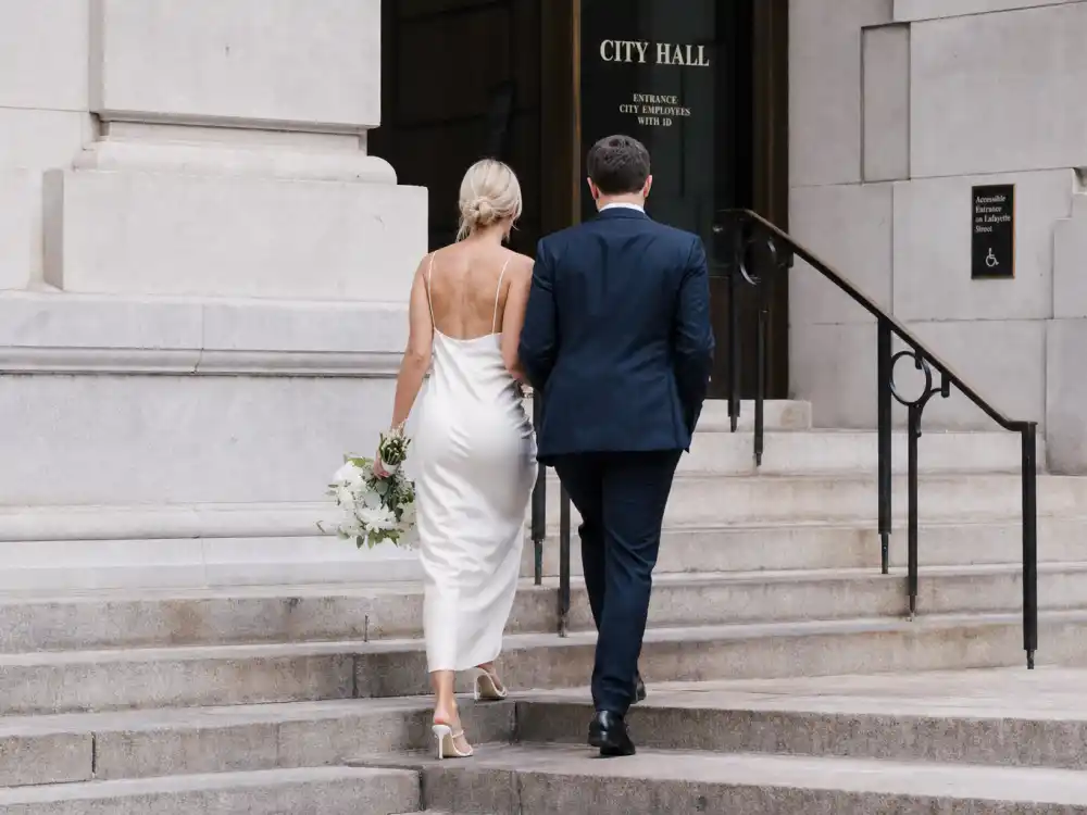 Minimal white sheath slip dress and navy suit celebrity courthouse wedding outfits on historic courthouse stone steps from behind