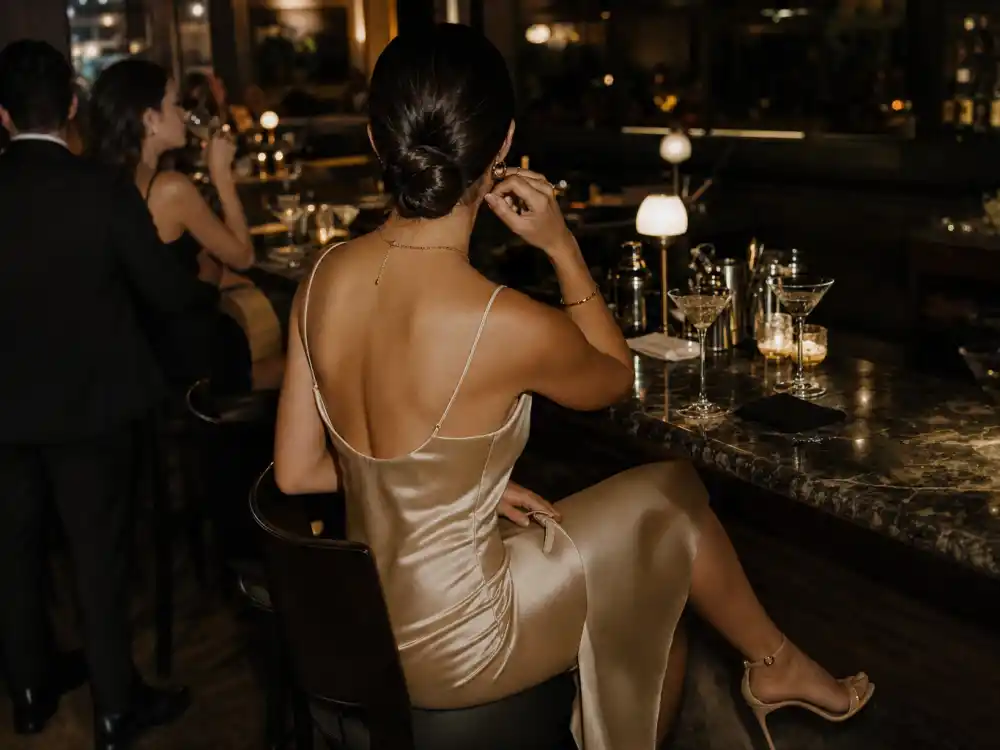 Cocktail bar celebrity date night outfit ideas with champagne satin slip dress and gold jewelry at upscale lounge counter