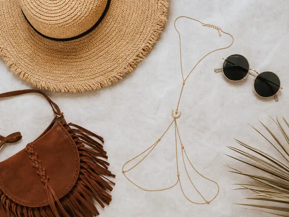 Round sunglasses wide-brim hat gold body chain and fringe crossbody bag flat lay finishing celebrity festival fashion summer 2026 accessory stacking rules