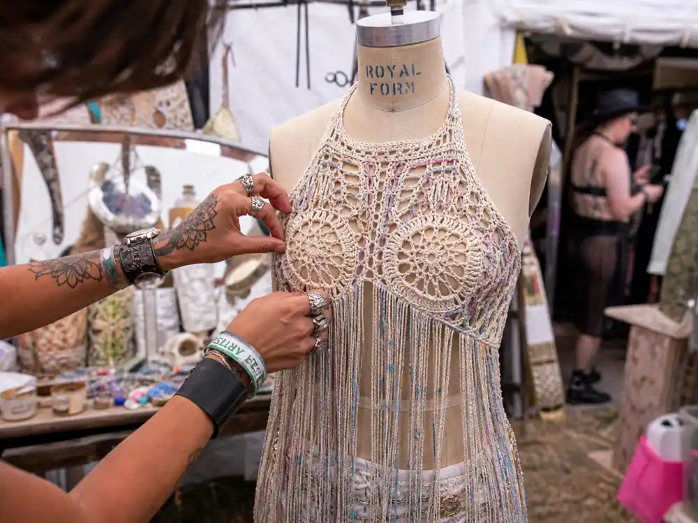 Stylist hands adjusting crochet fringe top on dress form backstage at summer music festival showing craftsmanship behind celebrity festival fashion summer 2026
