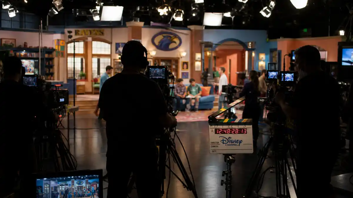 Celebrity kids who started on Disney Channel — TV soundstage from behind with anonymous crew silhouettes and Disney clapperboard editorial without identifiable faces