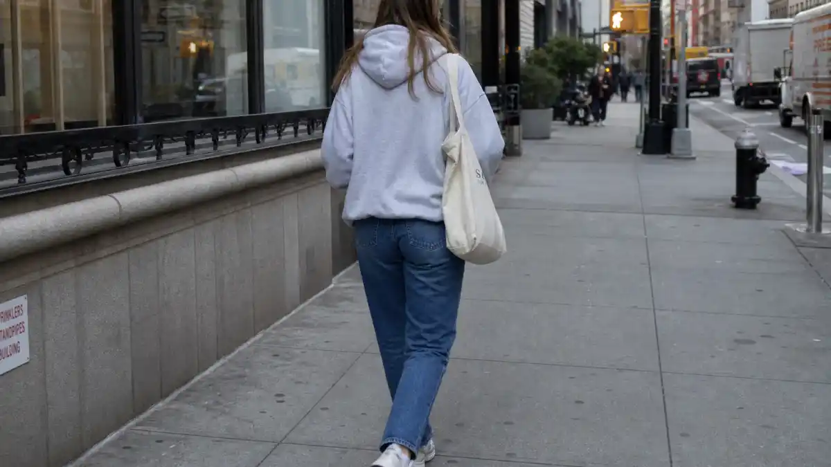Celebrity normcore fashion trend — anonymous woman from behind in gray hoodie, straight blue jeans, and white sneakers with canvas tote on city sidewalk