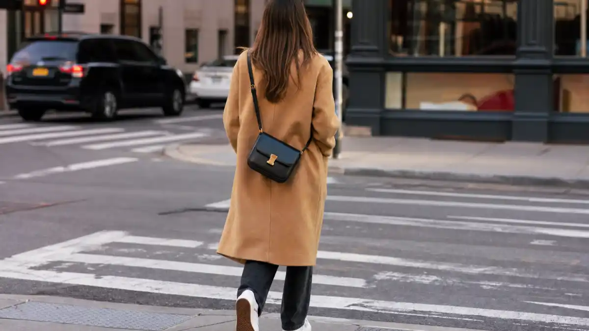 Celebrity off duty street style guide — camel coat straight jeans white sneakers and crossbody bag on NYC crosswalk seen from behind