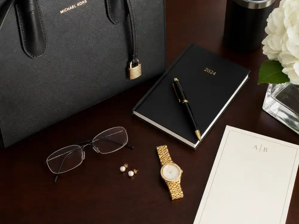 Thin-frame glasses pearl studs gold watch and structured leather tote for celebrity office chic outfit inspiration accessory detail on executive desk