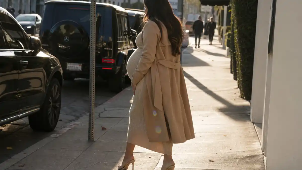 Celebrity pregnancy street style 2026 — anonymous pregnant woman in beige ribbed midi dress and camel trench coat walking Los Angeles sidewalk seen from behind