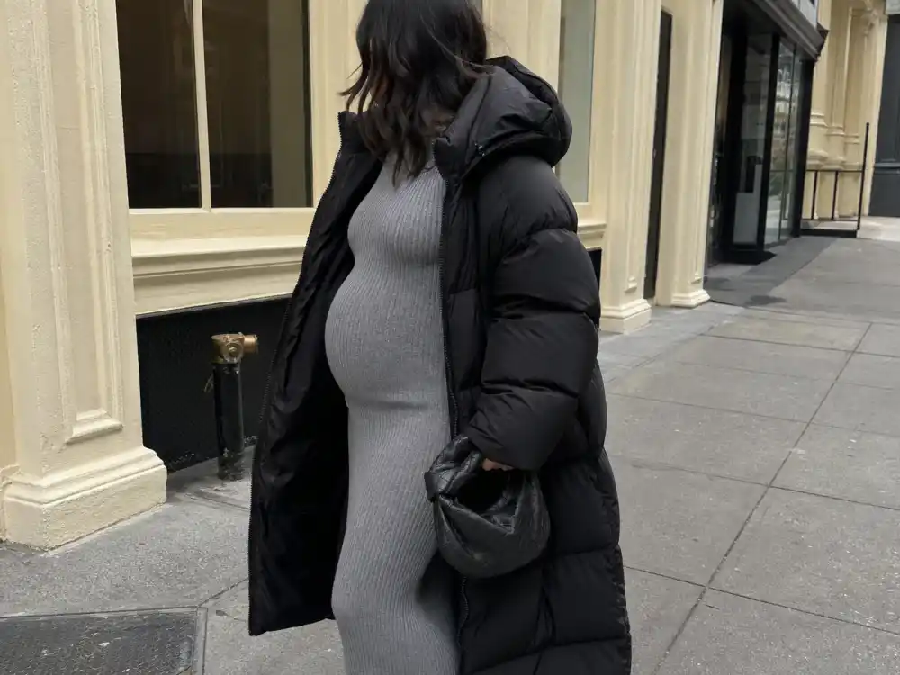 Anonymous pregnant woman in oversized black puffer coat over gray ribbed dress showing celebrity pregnancy street style 2026 layered outerwear formula