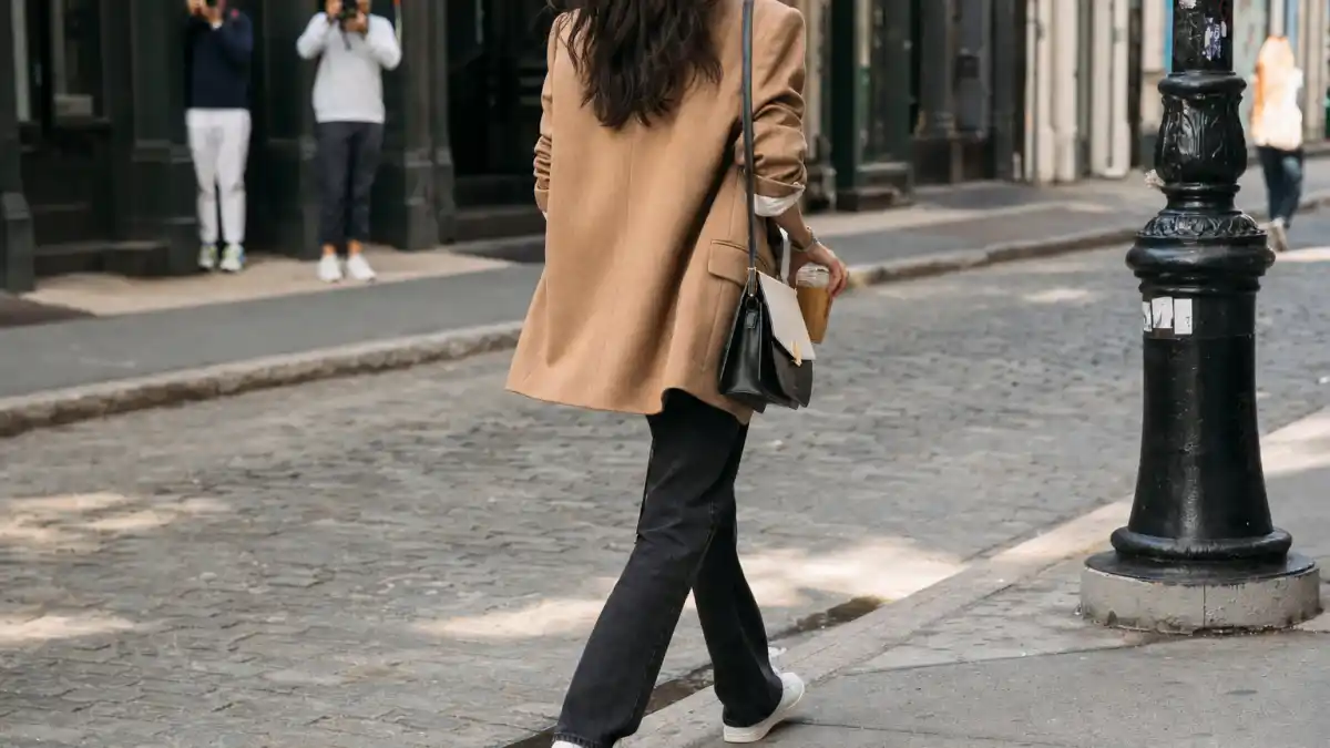 Celebrity street style — camel oversized blazer white tee black straight-leg jeans white sneakers and crossbody bag on SoHo cobblestone sidewalk seen from behind