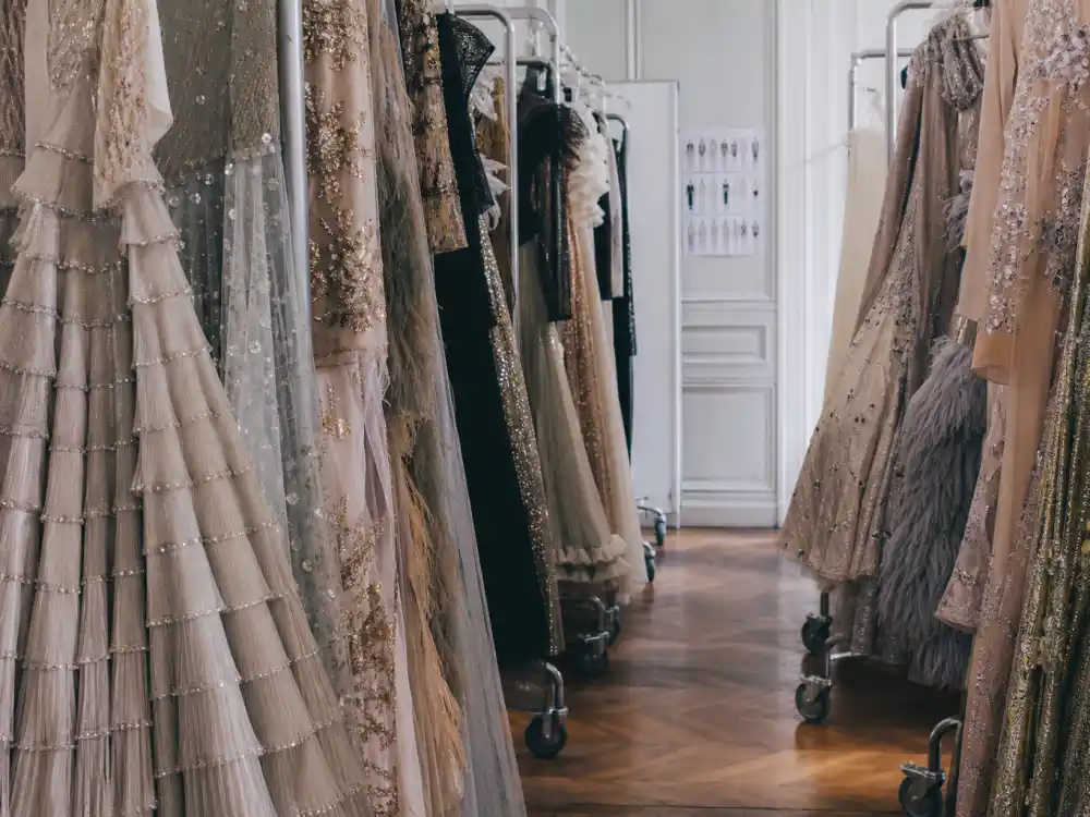 Haute couture evening gowns on rolling racks in designer showroom aisle showing luxury construction behind celebrity style icons of all time atelier preparation