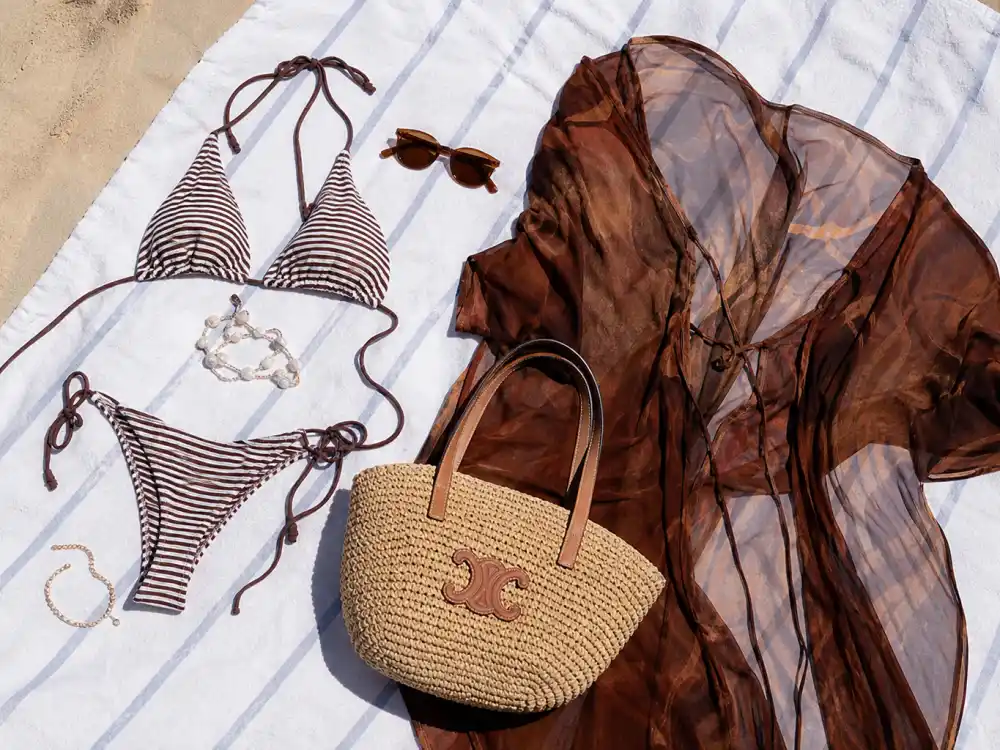 Striped bikini sheer beach cover-up and woven tote flat lay on sand illustrating celebrity summer vacation outfits 2026 beach style codes