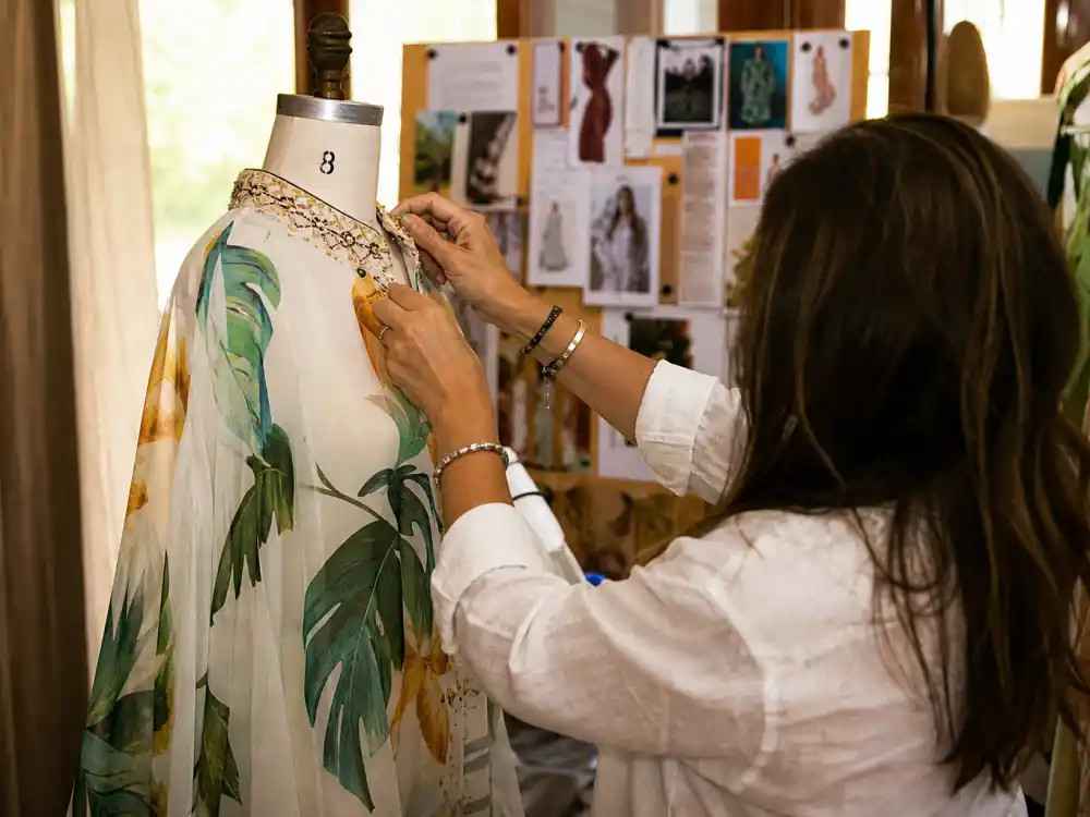 Stylist hands adjusting flowing resort caftan on dress form showing couture construction behind celebrity summer vacation outfits 2026 backstage craftsmanship