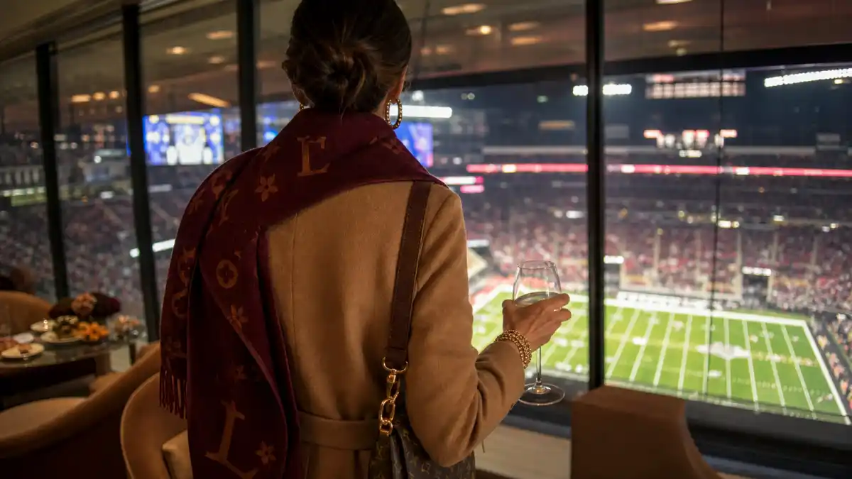 Celebrity super bowl fashion 2026 — anonymous woman from behind in camel cashmere coat and burgundy team-color scarf in NFL stadium luxury suite watching game day action