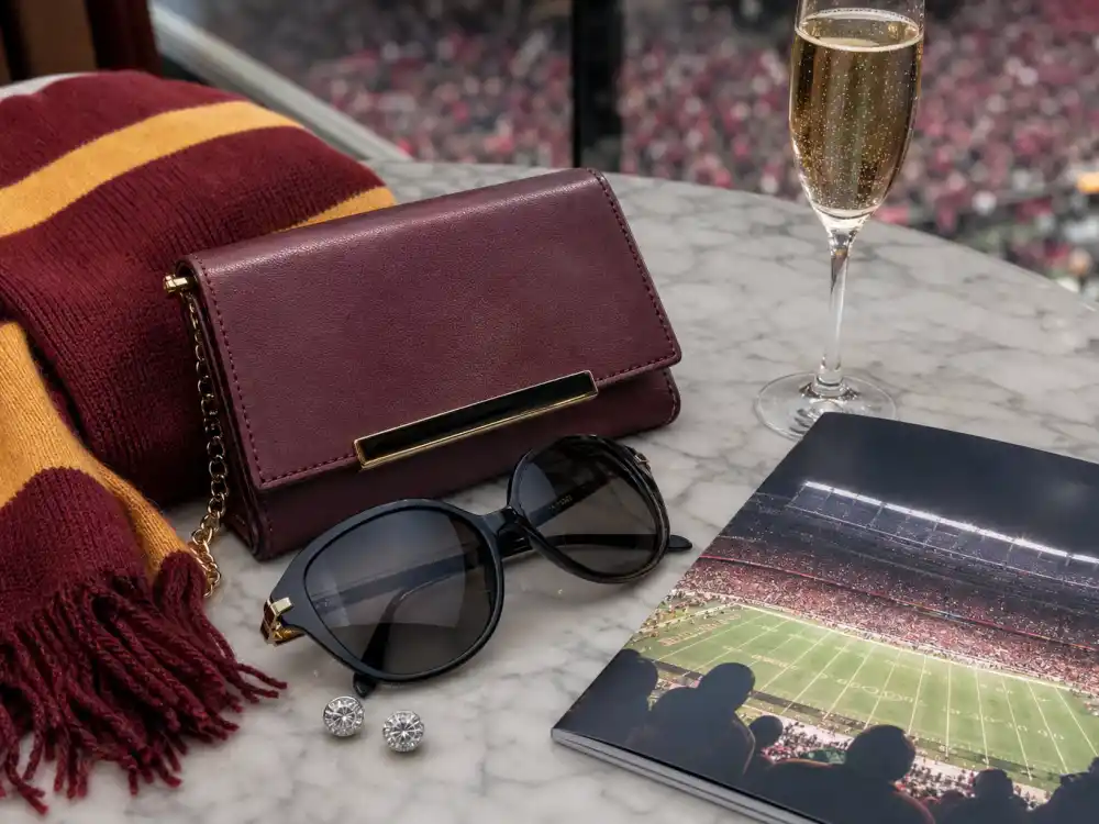 Luxury game day accessories flat lay with team-color cashmere scarf designer sunglasses champagne and leather clutch defining celebrity super bowl fashion 2026 suite styling