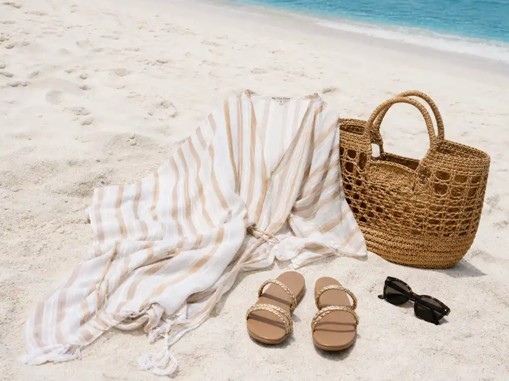 Striped resort cover-up gold sandals and woven tote flat lay on sand illustrating celebrity vacation resort wear beach style codes