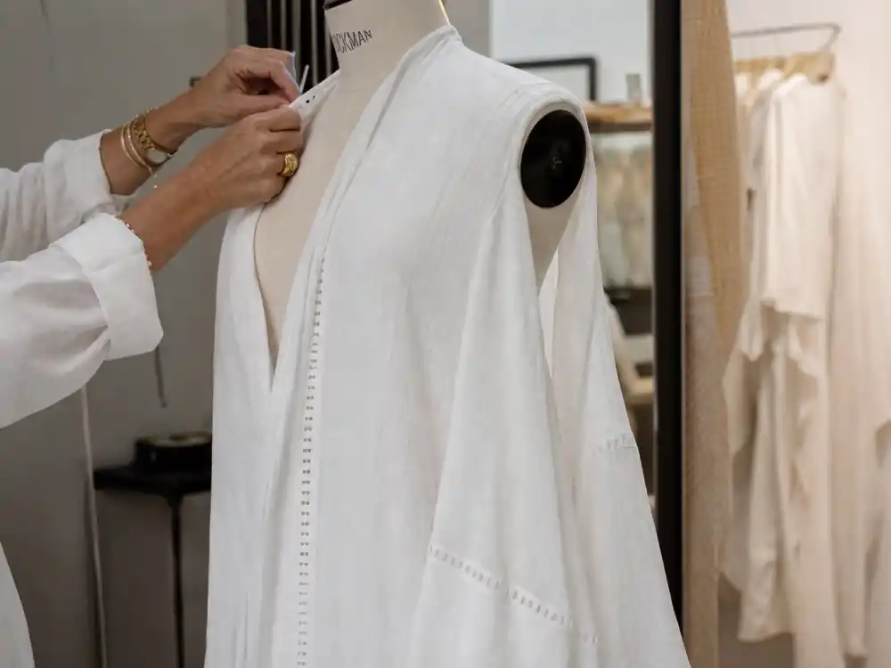 Stylist hands adjusting flowing resort caftan on dress form showing couture construction behind celebrity vacation resort wear backstage craftsmanship