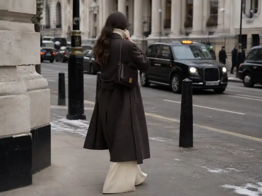 Anonymous woman from behind layering turtleneck sweater under long wool coat with wide leg trousers on cold city street illustrating celebrity winter coat style 2026 layering codes