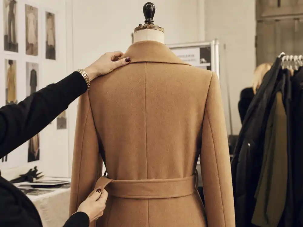Fashion stylist hands adjusting collar and belt on long camel wool coat on dress form showing backstage craftsmanship behind celebrity winter coat style 2026 tailoring