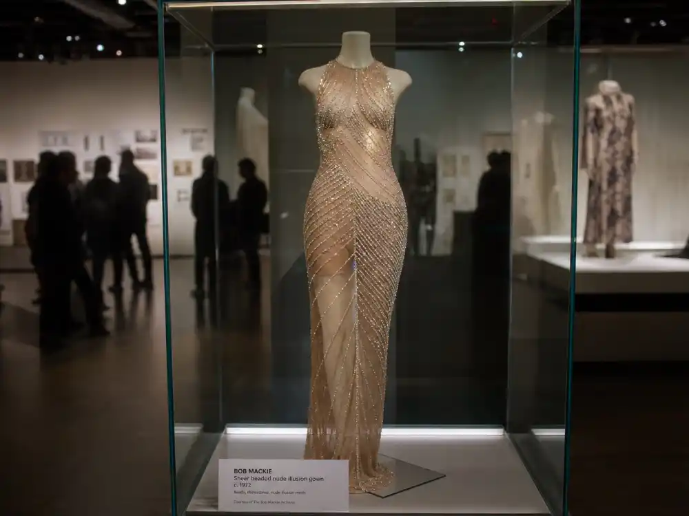 Fashion museum exhibition preserving Cher naked dress Bob Mackie history legacy — sheer beaded gown on gallery mannequin behind glass
