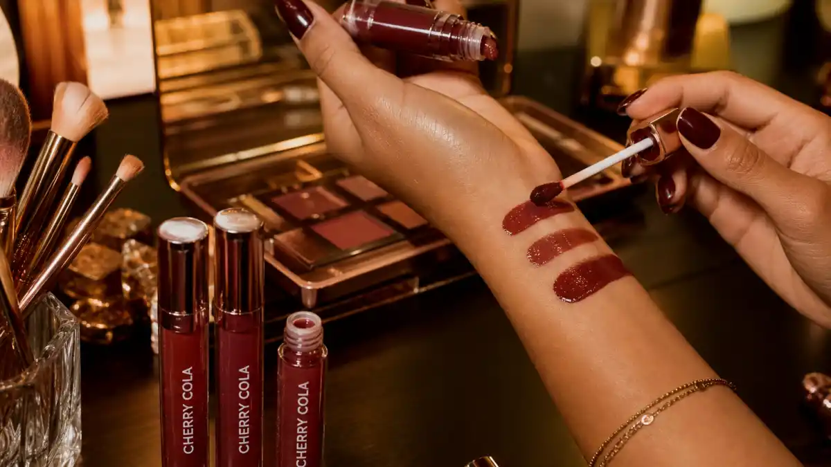 Cherry cola beauty celebrity trend — hands swatching glossy burgundy lip gloss on arm at warm-lit vanity with cherry cola lip tubes and bronze eyeshadow palette