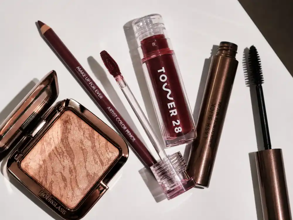 Flat lay of cherry cola lip liner, glossy cola lip oil, and bronze highlighter products mapping cherry cola beauty celebrity trend shopping essentials