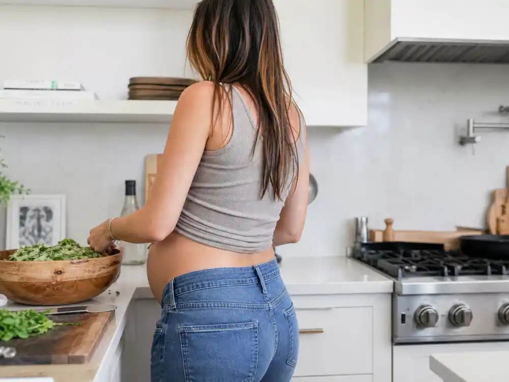 Pregnant woman in casual ribbed tank and jeans in modern kitchen illustrating Chrissy Teigen maternity style outfits off-duty at-home pregnancy fashion