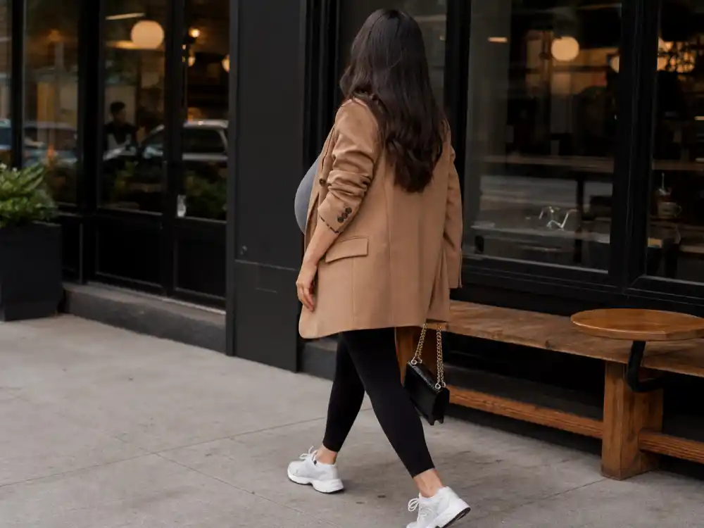 Pregnant woman in oversized camel blazer leggings and sneakers outside coffee shop for Chrissy Teigen maternity style outfits street pregnancy uniform