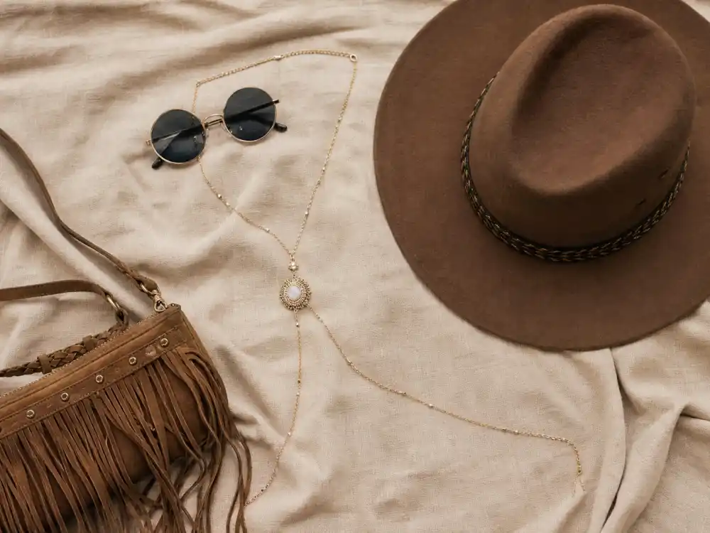 Round sunglasses wide-brim hat gold body chain and fringe crossbody bag flat lay finishing coachella celebrity outfits 2026 accessory stacking rules