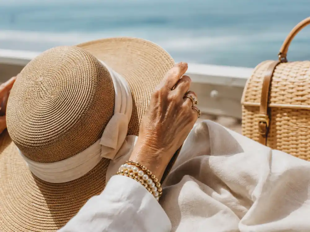 Hands adjusting wide-brim straw hat beside cream linen and wicker bag defining coastal grandmother celebrity style accessory codes stylists repeat