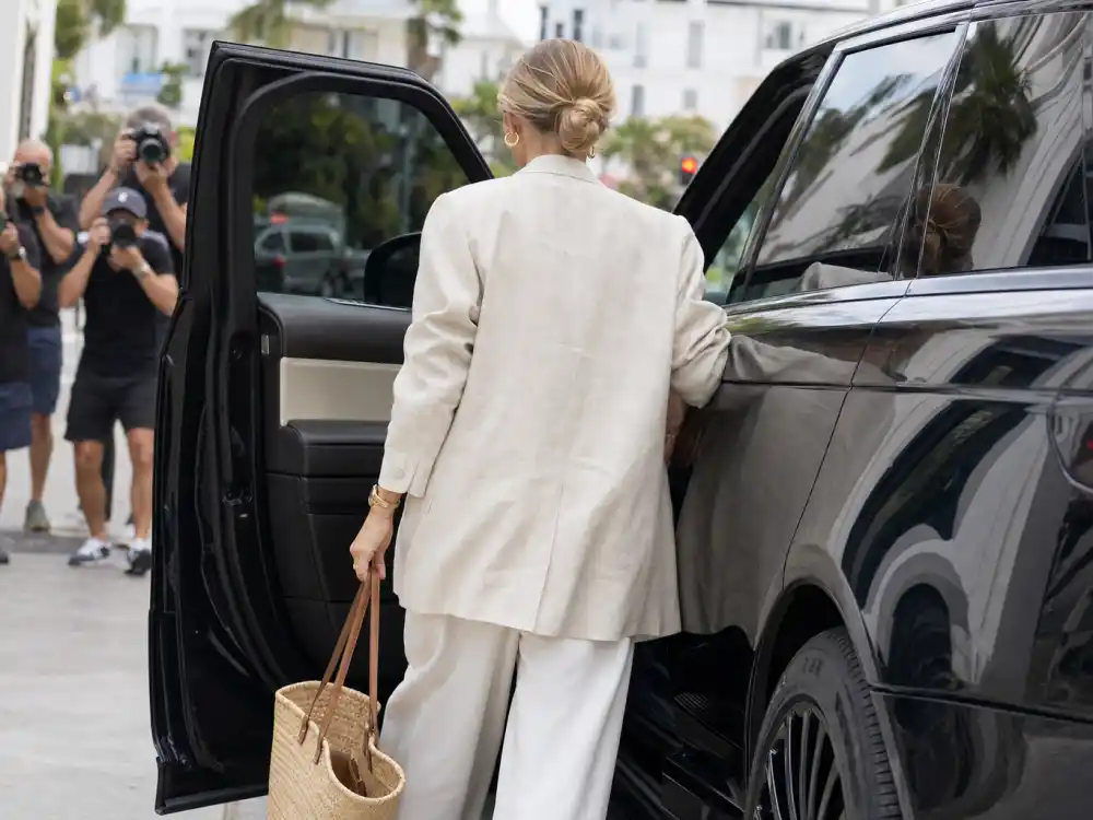 Anonymous woman from behind in coastal grandmother celebrity style cream linen blazer white trousers and straw tote exiting car on upscale coastal sidewalk