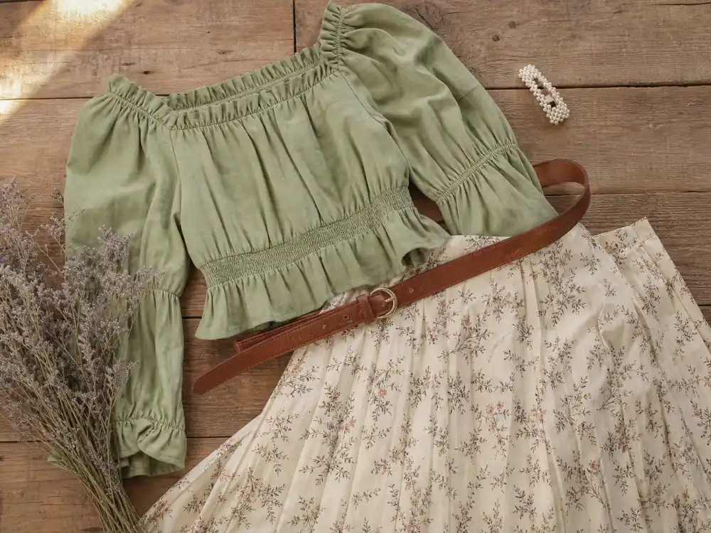 Cottagecore celebrity fashion looks flat lay with sage puff-sleeve blouse cream floral midi skirt brown leather belt and dried lavender on rustic wood