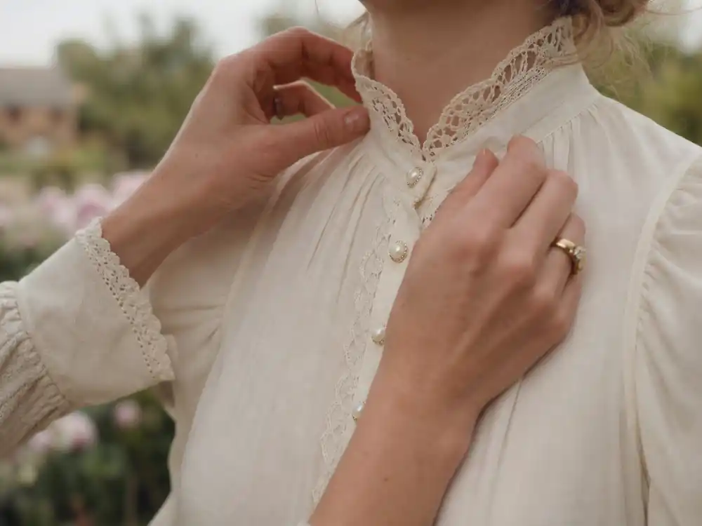 Hands adjusting vintage lace collar on cream puff-sleeve blouse defining cottagecore celebrity fashion looks romantic styling detail with pearl buttons