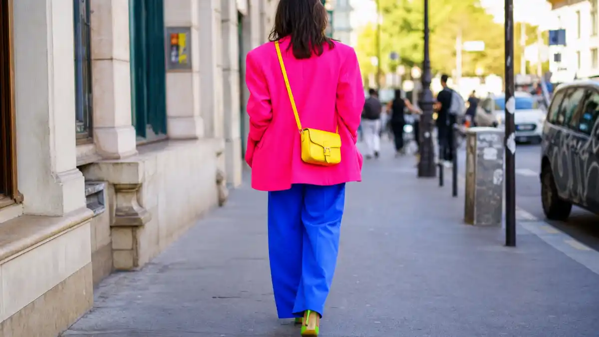 Dopamine dressing celebrity outfits — anonymous woman from behind in hot pink blazer electric blue trousers and sunny yellow bag on colorful city sidewalk