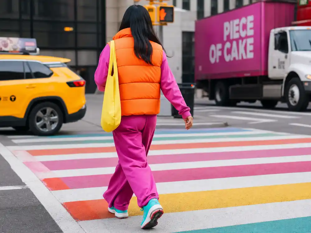 Anonymous person crossing urban crosswalk from behind in dopamine dressing celebrity outfits tangerine vest magenta pants turquoise sneakers lemon tote