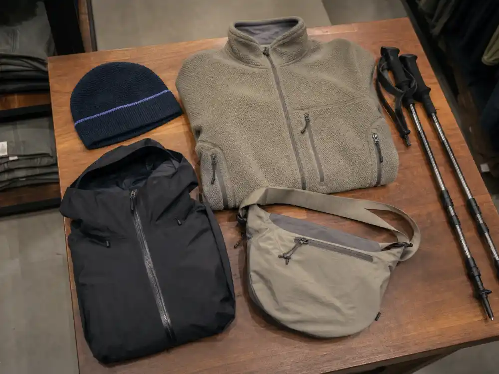 Retail flat lay of fleece pullover, technical shell, beanie, and nylon crossbody mapping gorpcore celebrity street style shopping essentials